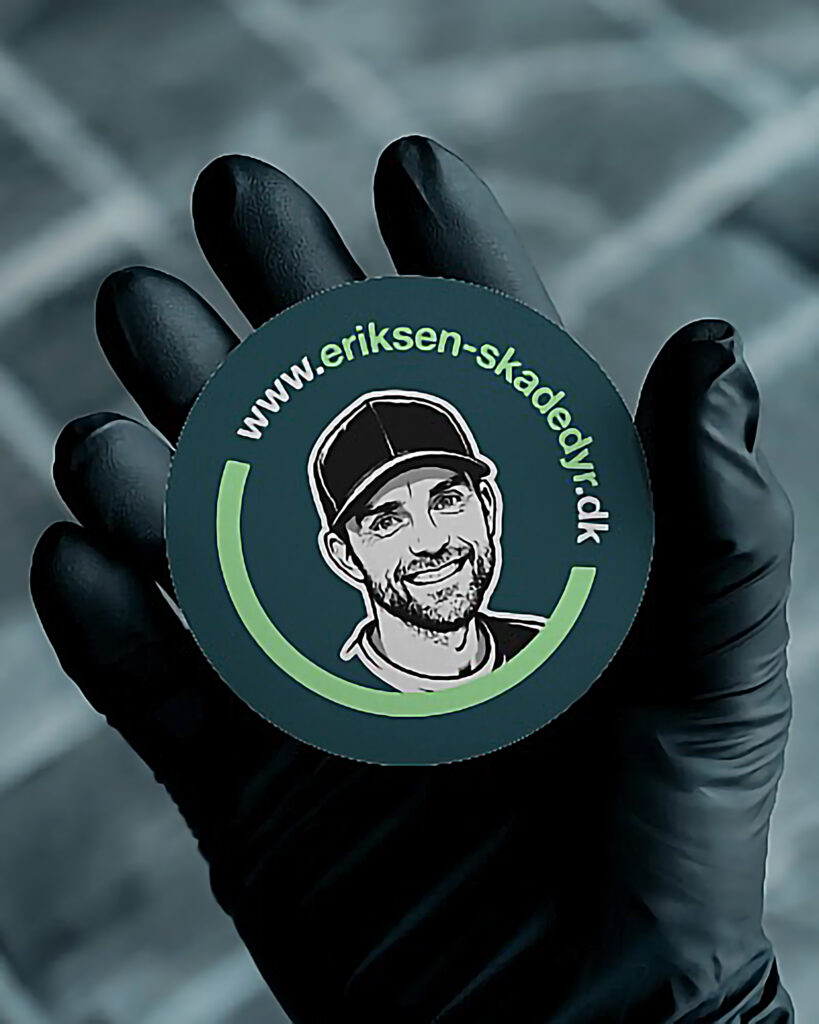 Visitkort Eriksen Skadedyrsservice sort latexhandske foran uskarpt grøn-hvis ternet baggrund holder et rund visitkort af Eriksen skadedyrsservice med ansigt af Mads Eriksen og www.eriksen-skadedyr.dk websitet på visitkortet